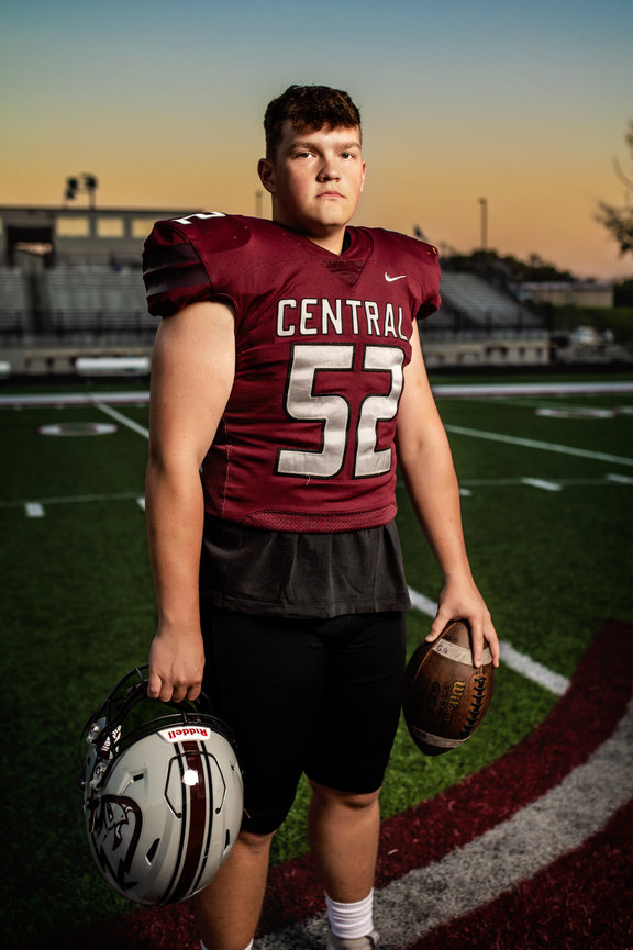 Westosha Central football player in uniform stands on a field at sunset holding a helmet and football for his senior pictures.