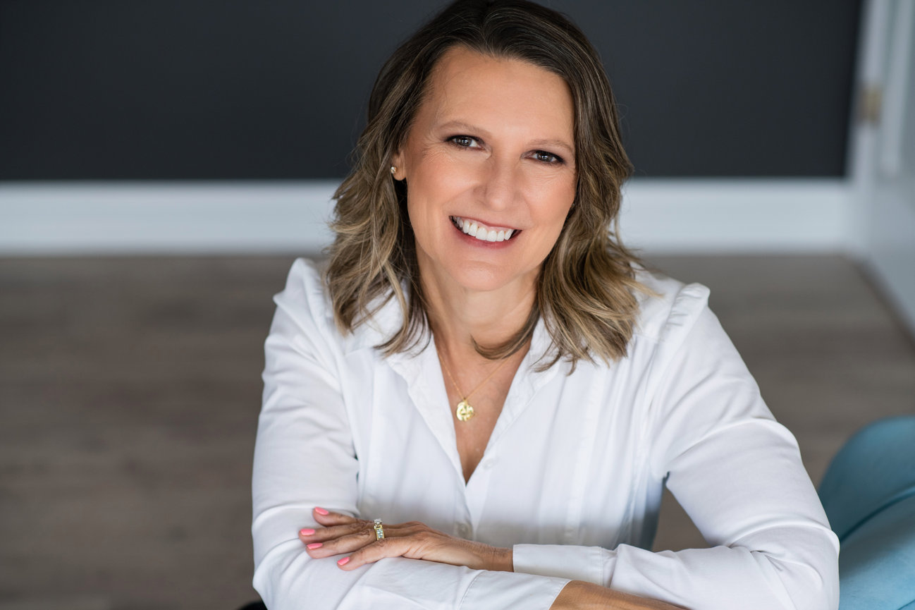 Smiling woman in a white blouse, seated with arms crossed, against a dark background for her branding session in a Dublin Ohio boutique studio.