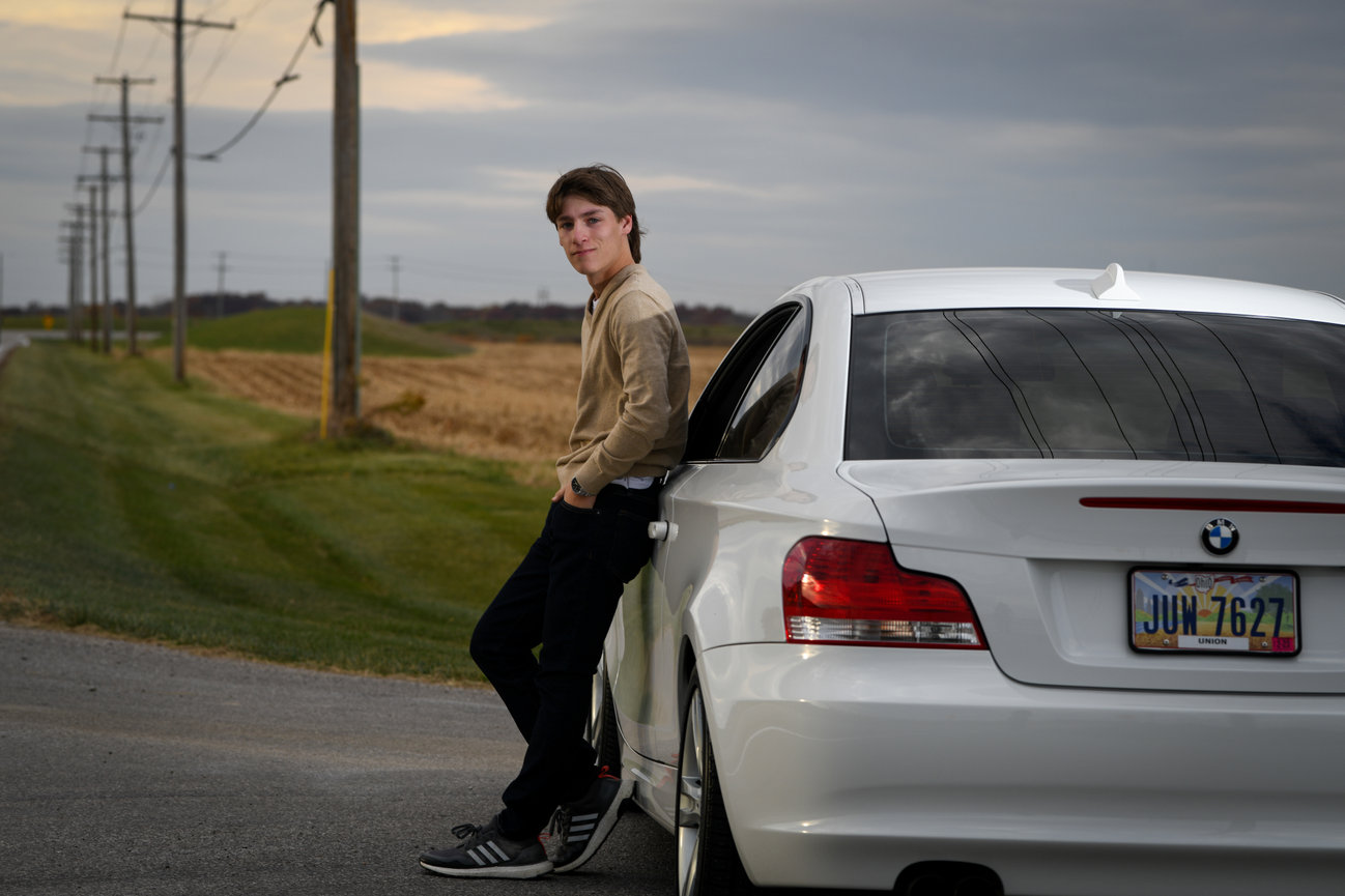 Dark haired teenage boy leaning against his white BMW on a rural road with clouds in Dublin OH.