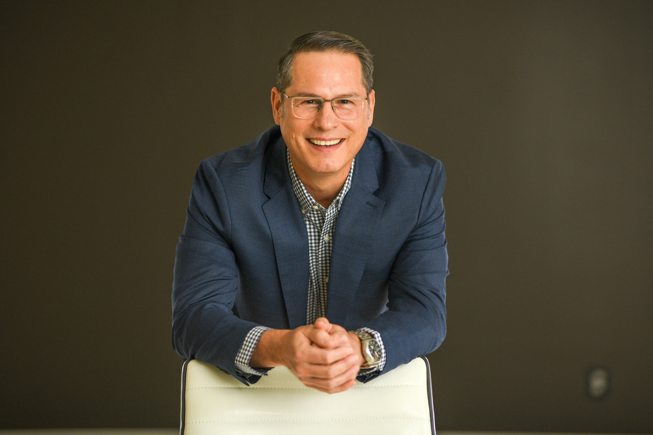 Smiling man in a blue blazer leaning on a chair, against a neutral background. Headshot Photographer Dublin Ohio