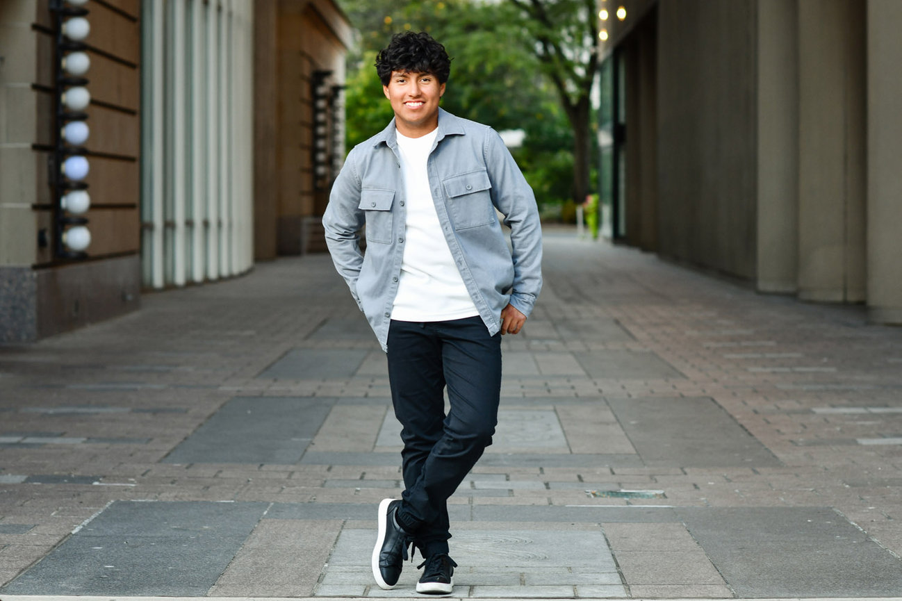 Dark haired teenage boy standing in an urban setting wearing jeans and an unbuttoned jean shirt.