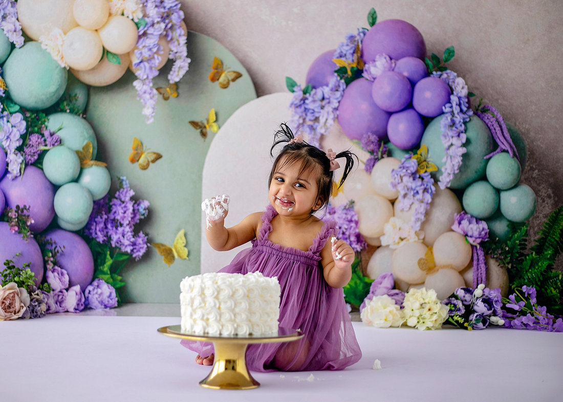 Smiling baby girl in a purple dress enjoying cake during a butterfly and lavender-themed cake smash with pastel balloons and flowers by Cake Smash by Chris in Point Cook.
