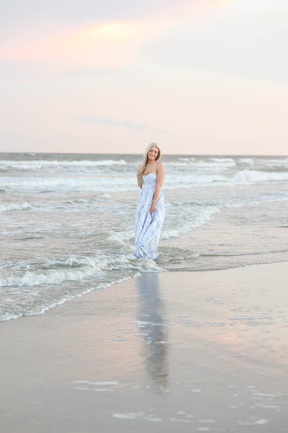 Woman in a flowing dress stands at the edge of the ocean during a pastel sunset.